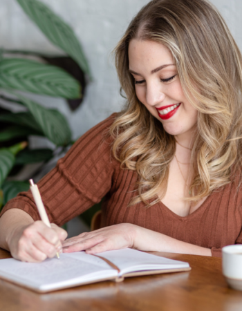 Woman smiling while writing in a notebook at a desk — how to write an about page that connects with clients
