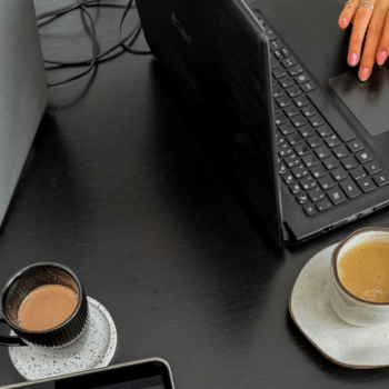 Overhead view of a dark desk with two laptops, a hand on a trackpad and two cups of coffee, the setup for planning Black Friday emails