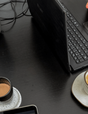 Overhead view of a dark desk with two laptops, a hand on a trackpad and two cups of coffee, the setup for planning Black Friday emails
